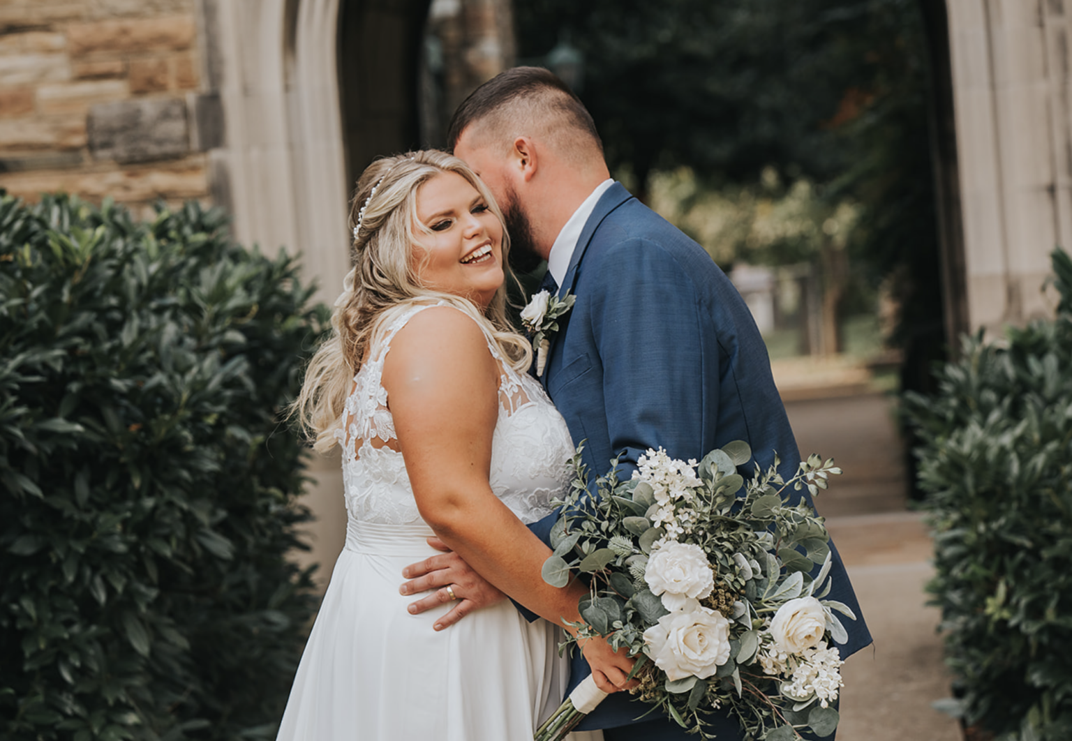 Elopement Wedding A bride and groom share a joyous moment on their wedding day. The bride, in a white lace dress, smiles brightly while holding a bouquet of white roses and greenery. The groom, in a blue suit, is nuzzling close to her. They stand in front of a stone archway surrounded by greenery. Elopements Inc